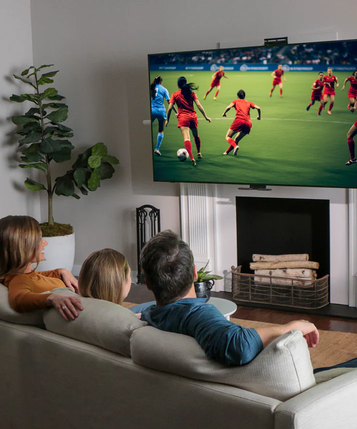 Family watching soccer on a wall-mounted TV above fireplace in a modern living room
